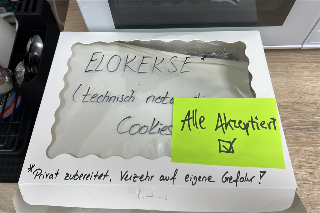 Foto einer geöffneten weißen Keksbox mit handbeschriftetem Zettel „ELOKERSE (technisch notwendige Cookies)“ und gelbem Post-it mit der Aufschrift „Alle Akzeptiert“ und einem Häkchen. Die Box steht auf einem Bürotisch neben einer Kaffeemaschine – Symbol für Teamgeist und kreative IT-Kultur.